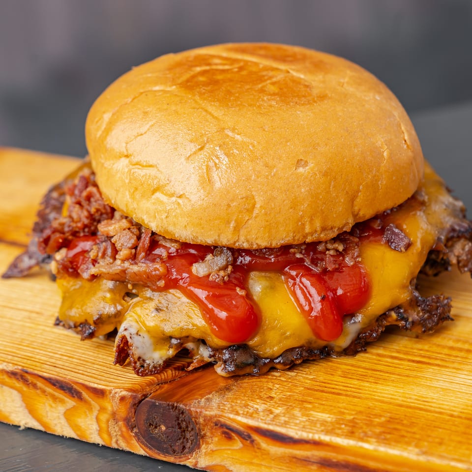 Close-up cheeseburger topped with bacon, ketchup, pickles, and melted cheese on a wooden serving board.