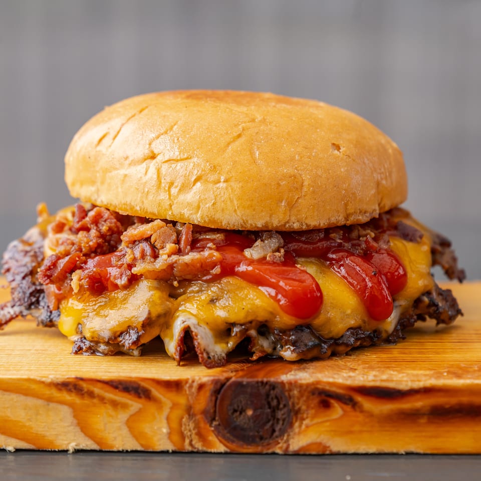 Close-up of a loaded cheeseburger with bacon, vegetables, and ketchup on a wooden cutting board.