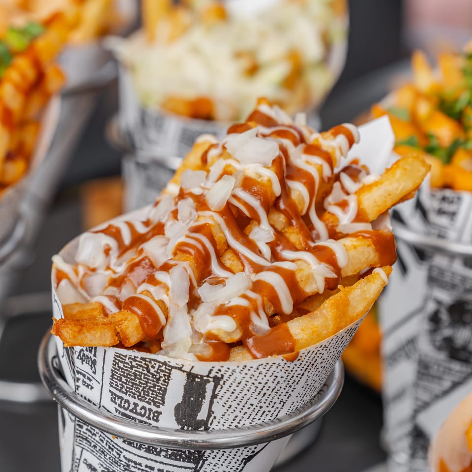 Crispy fries in a newspaper-lined cup topped with red and white sauces and chopped onions, surrounded by other loaded fries.