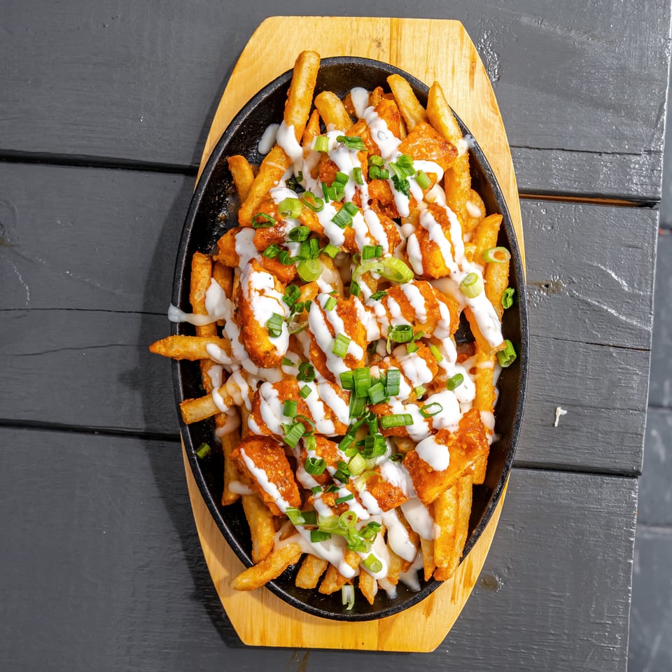 Loaded fries topped with crispy chicken, orange sauce, white drizzle, and chopped green onions in a cast-iron skillet.