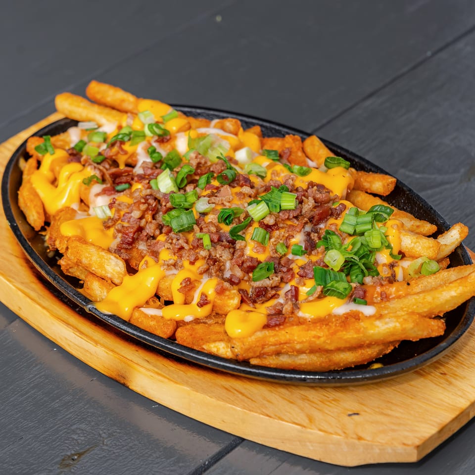 Loaded fries topped with melted cheese, bacon, ground meat, and green onions, served in a black skillet on a wooden board.
