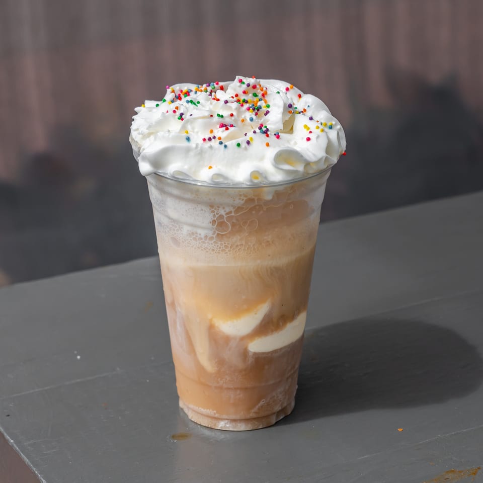 Iced coffee drink topped with whipped cream and colorful sprinkles in a clear plastic cup.