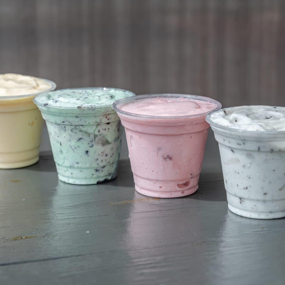 Four colorful cups of creamy frozen dessert, including vanilla, mint, pink berry, and cookies-and-cream flavors, arranged on a table.