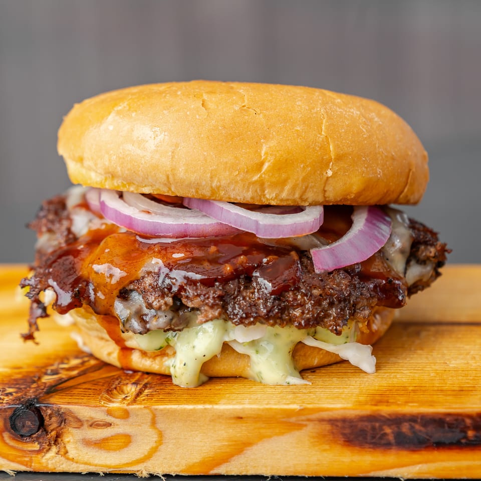 Gourmet cheeseburger with barbecue sauce, red onion, lettuce, and creamy dressing on a wooden board.