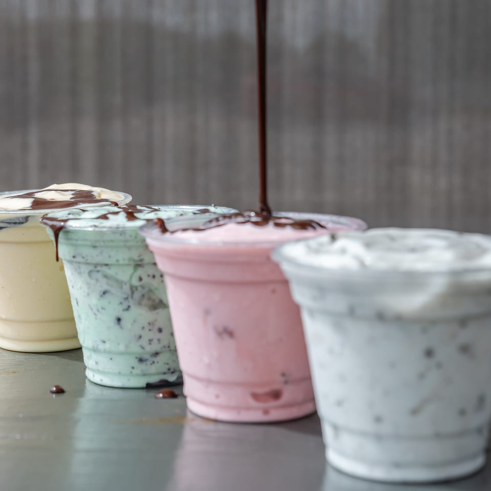 Four colorful ice cream cups topped with whipped cream and chocolate syrup sit on a metal table.