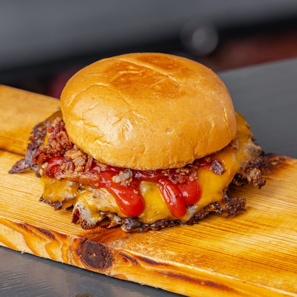 Loaded cheeseburger with bacon, shredded meat, tomato, and ketchup on a rustic wooden serving board.