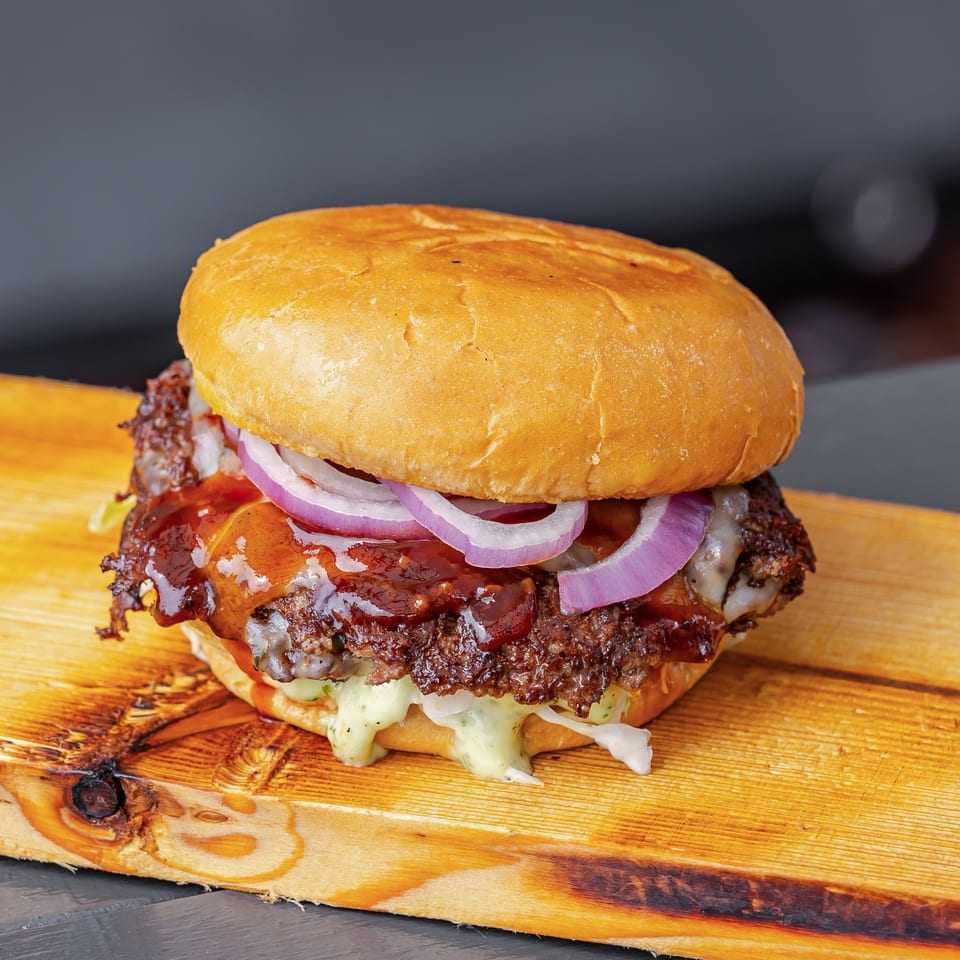 Close-up cheeseburger with a toasted bun, barbecue sauce, grilled beef patty, melted cheese, red onions, and creamy sauce on a wooden board.