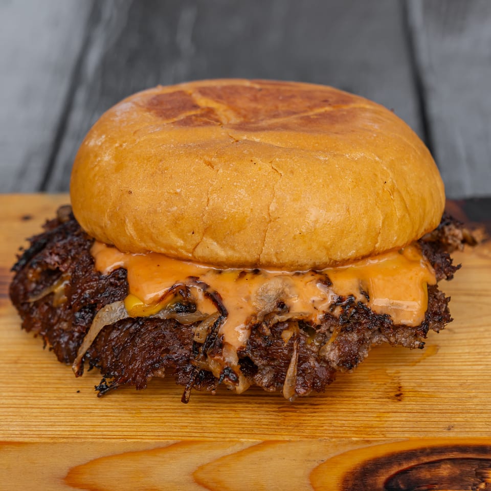 Close-up of a double cheeseburger with a toasted bun, melted cheese, grilled onions, sauce, and charred beef patties on a wooden board.