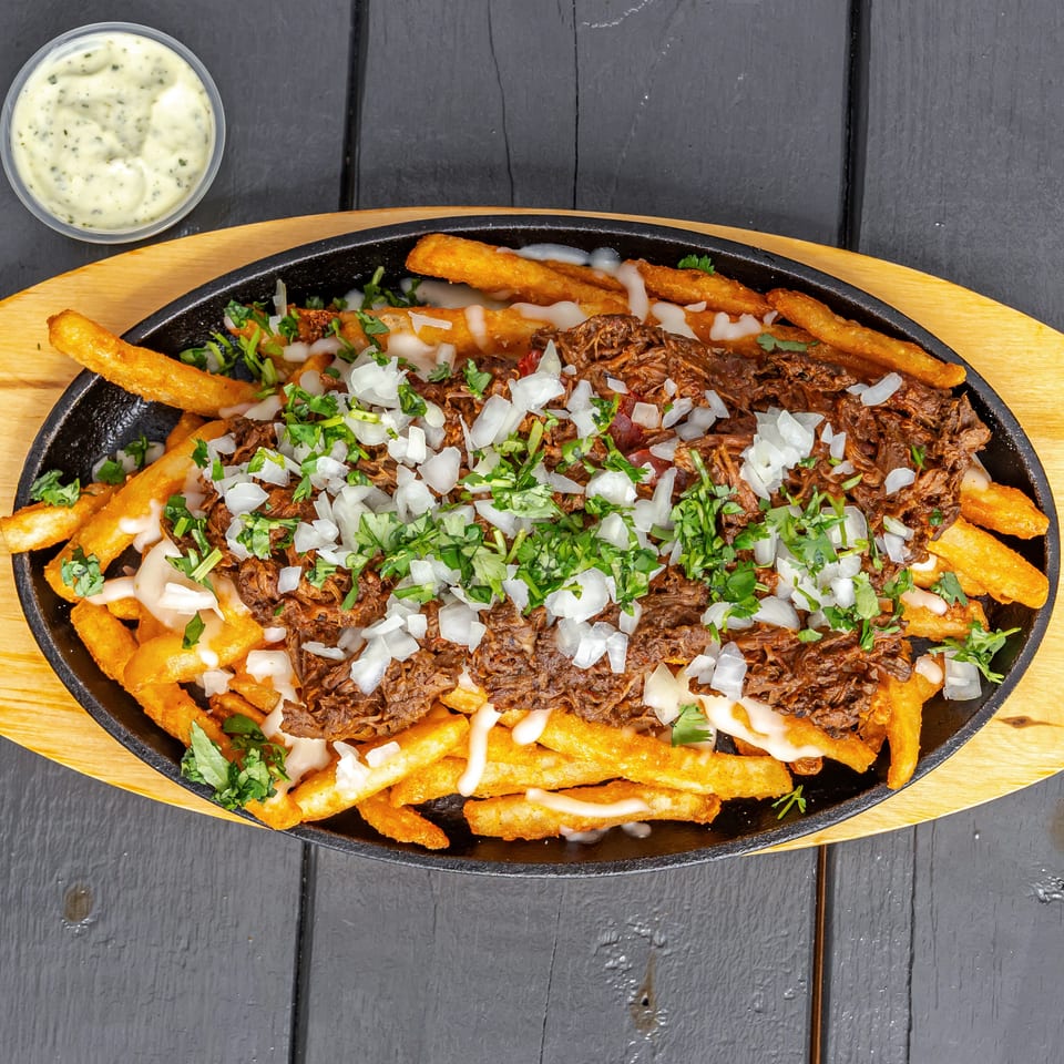 Loaded fries topped with shredded meat, white sauce, chopped onions, and cilantro in a cast-iron serving dish.