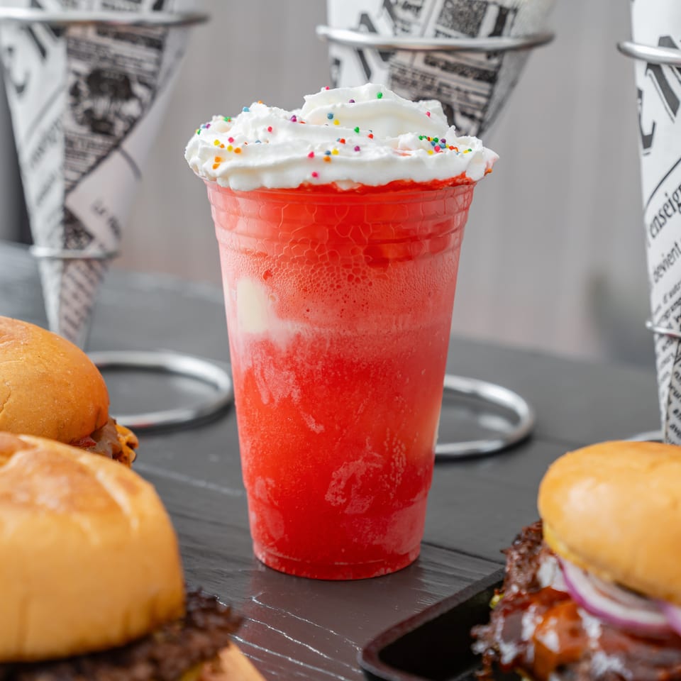 Bright red frozen drink topped with whipped cream and colorful sprinkles, surrounded by burgers on a restaurant table.