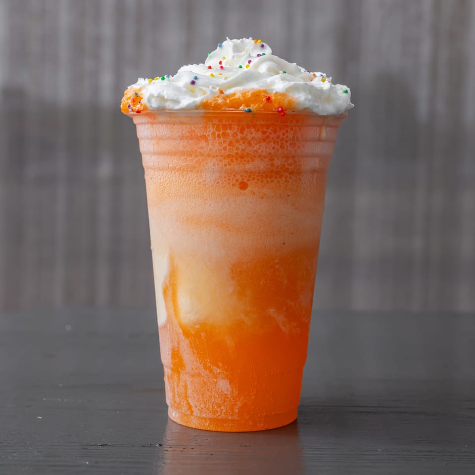 Orange frozen drink in a plastic cup topped with whipped cream and colorful sprinkles on a dark wooden table.