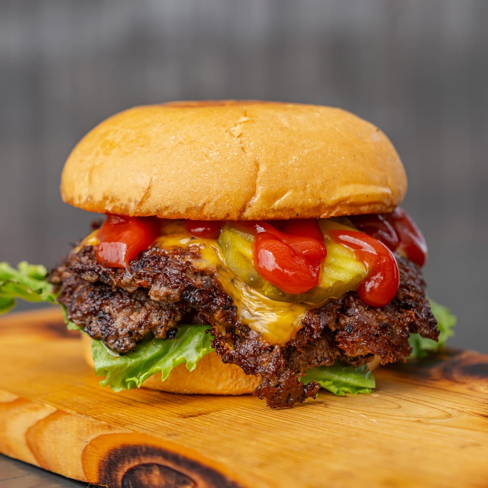 Close-up cheeseburger with a toasted bun, double beef patties, lettuce, pickles, tomatoes, and ketchup on a wooden board.