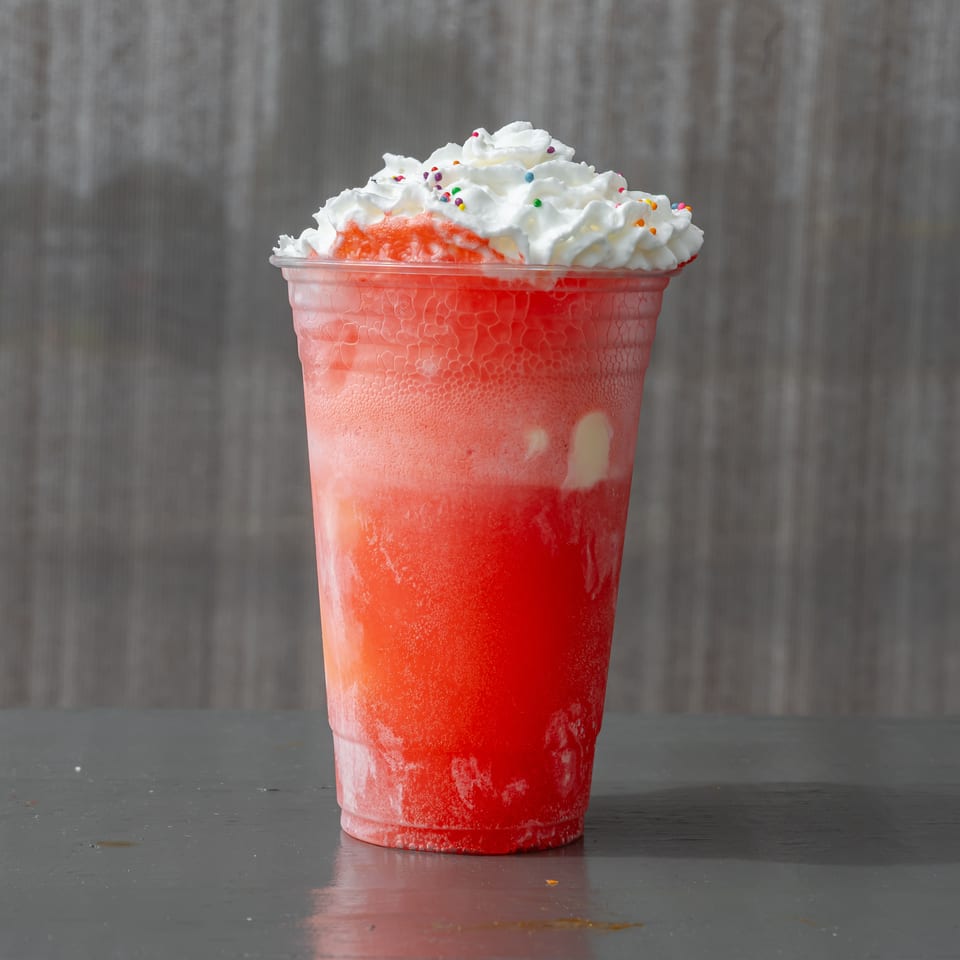 Red frozen beverage in a clear cup topped with whipped cream and colorful sprinkles on a tabletop.