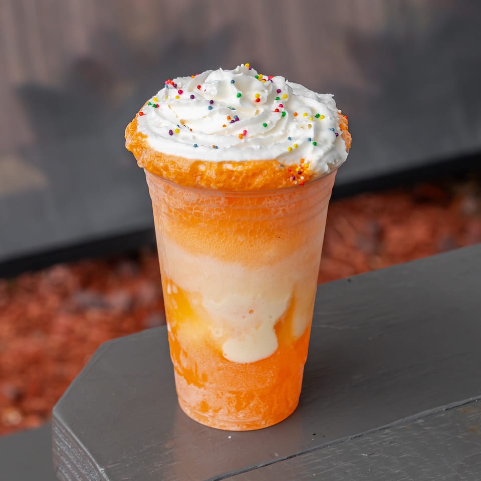 Orange creamy dessert drink topped with whipped cream and colorful sprinkles, served in a clear cup on a dark wooden table.
